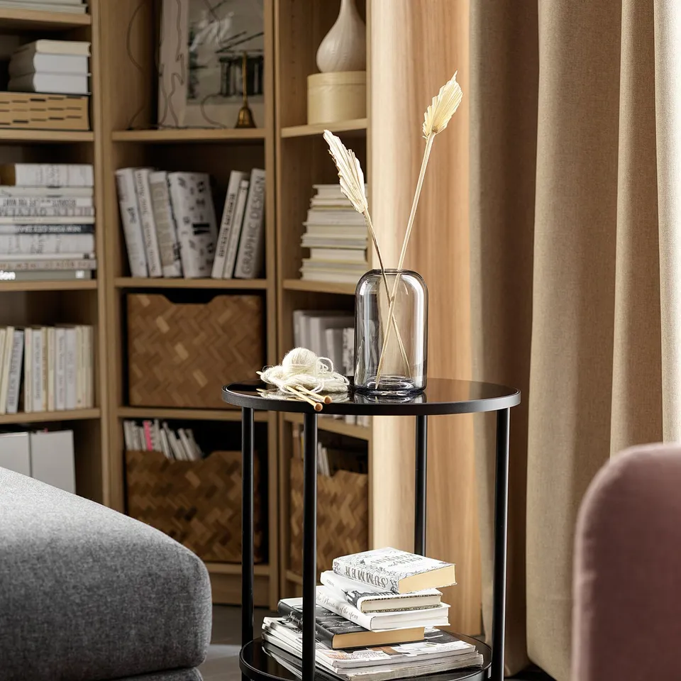 Modern living room with black ÄSPERÖD side table featuring smoky glass top, golden accents, and dried flowers in a clear vase.