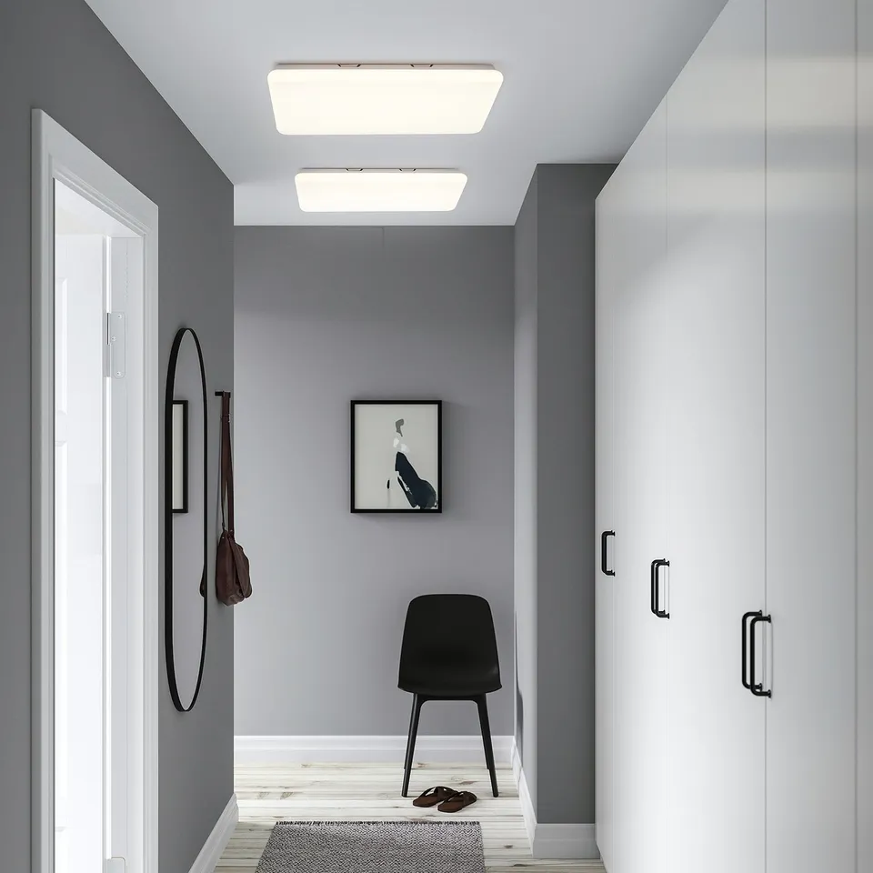 Modern hallway with white JETSTRÖM light panels, grey walls, black chair, and mirror. Panels are large and rectangular, emitting bright, even light.