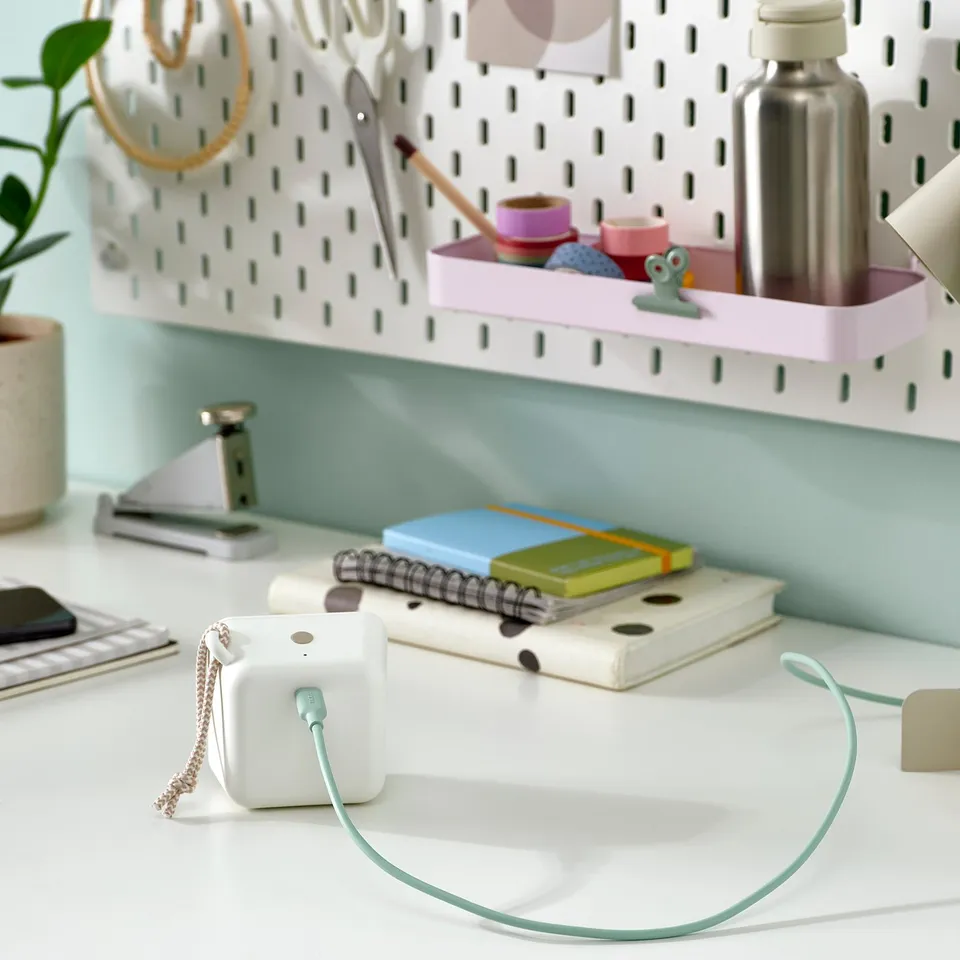 A green usb-c cable connects a small device on a white desk. The desk has a pegboard with various items including notebooks and a plant.