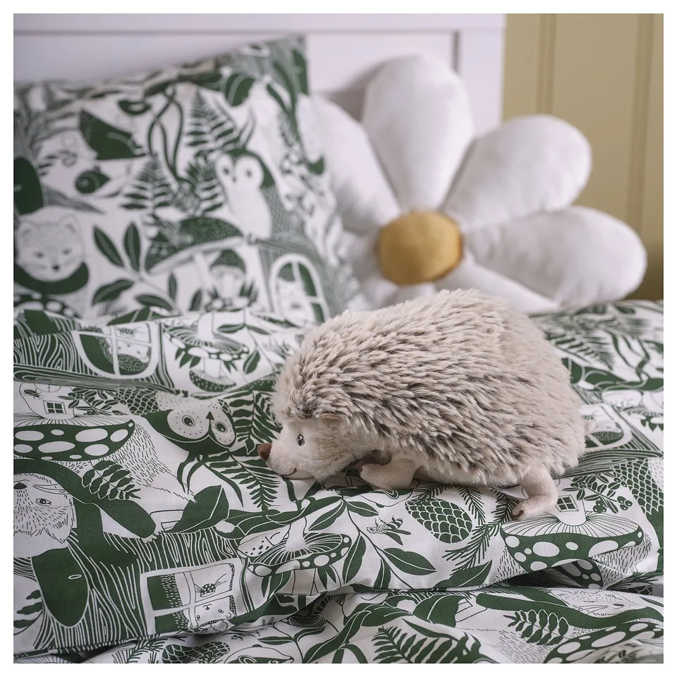 Hedgehog plush on forest bedding. Cream hedgehog, large white flower pillow, green/white pattern.