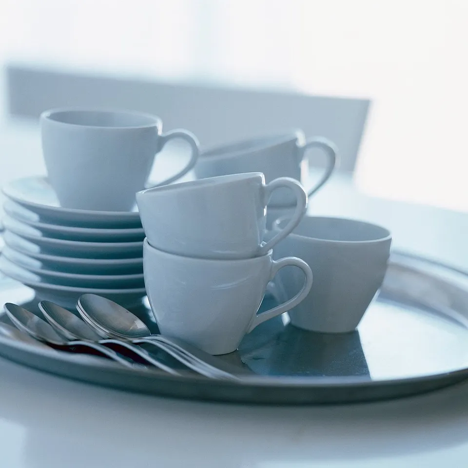 Stack of white VÄRDERA cups and plates on a tray, made of feldspar porcelain, square shape with soft lines.