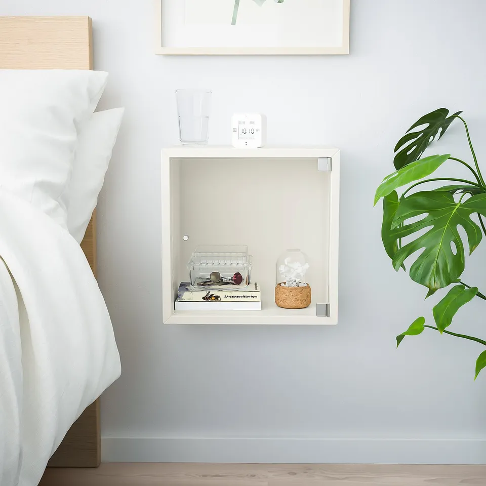 Small white wall-mounted ikea EKET cube shelf beside bed, holding glass jar, books, and plant.
