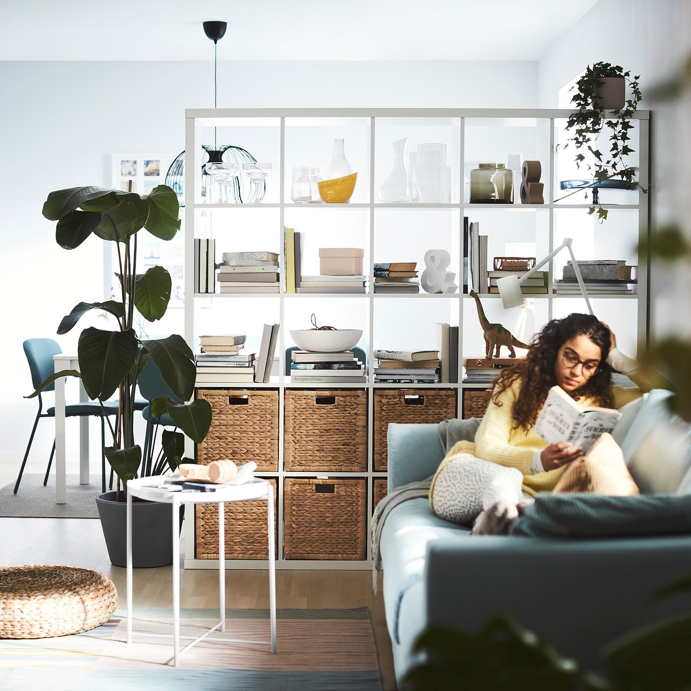 Person reading book on comfy blue couch beside KALLAX white shelving unit with wicker baskets, books, and plants.