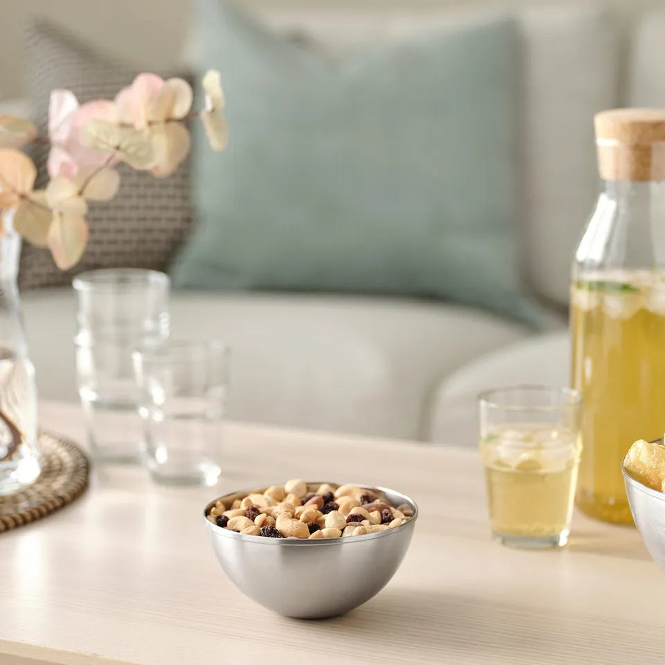 Silver stainless steel BLANDA BLANK bowl with snacks on table beside yellow drink. Smaller than glass.