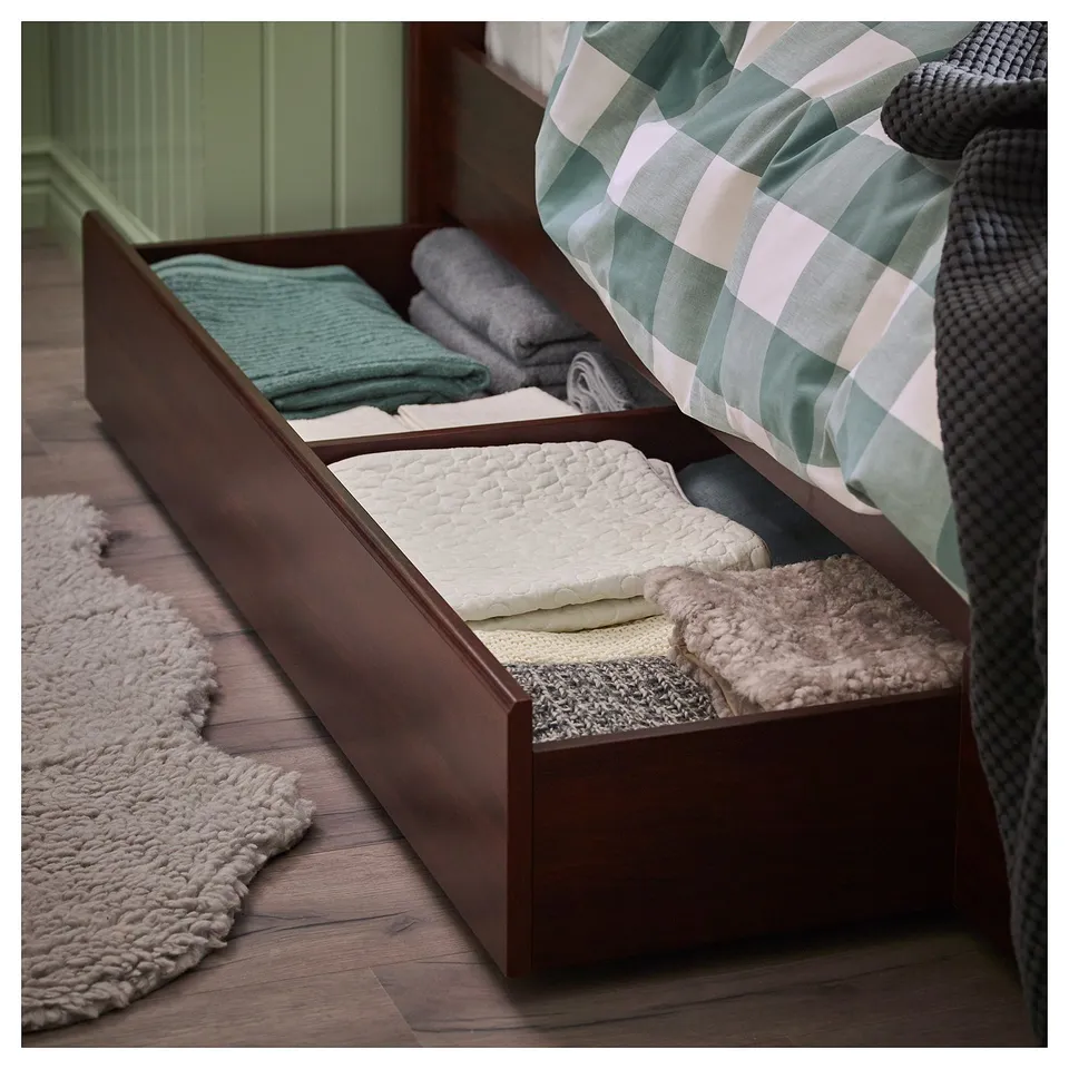 Wooden bed frame with under-storage. Folded blankets in turquoise, white, and grey. Plaid bedspread, grey knitted blanket. Soft rug on floor.