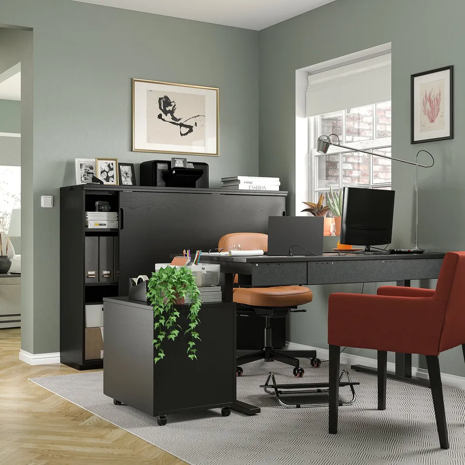 Home office scene: black desk, red server, printer, brown chair, potted plant, 2 red chairs, grey rug, green wall art, window, lamp, computer screen.