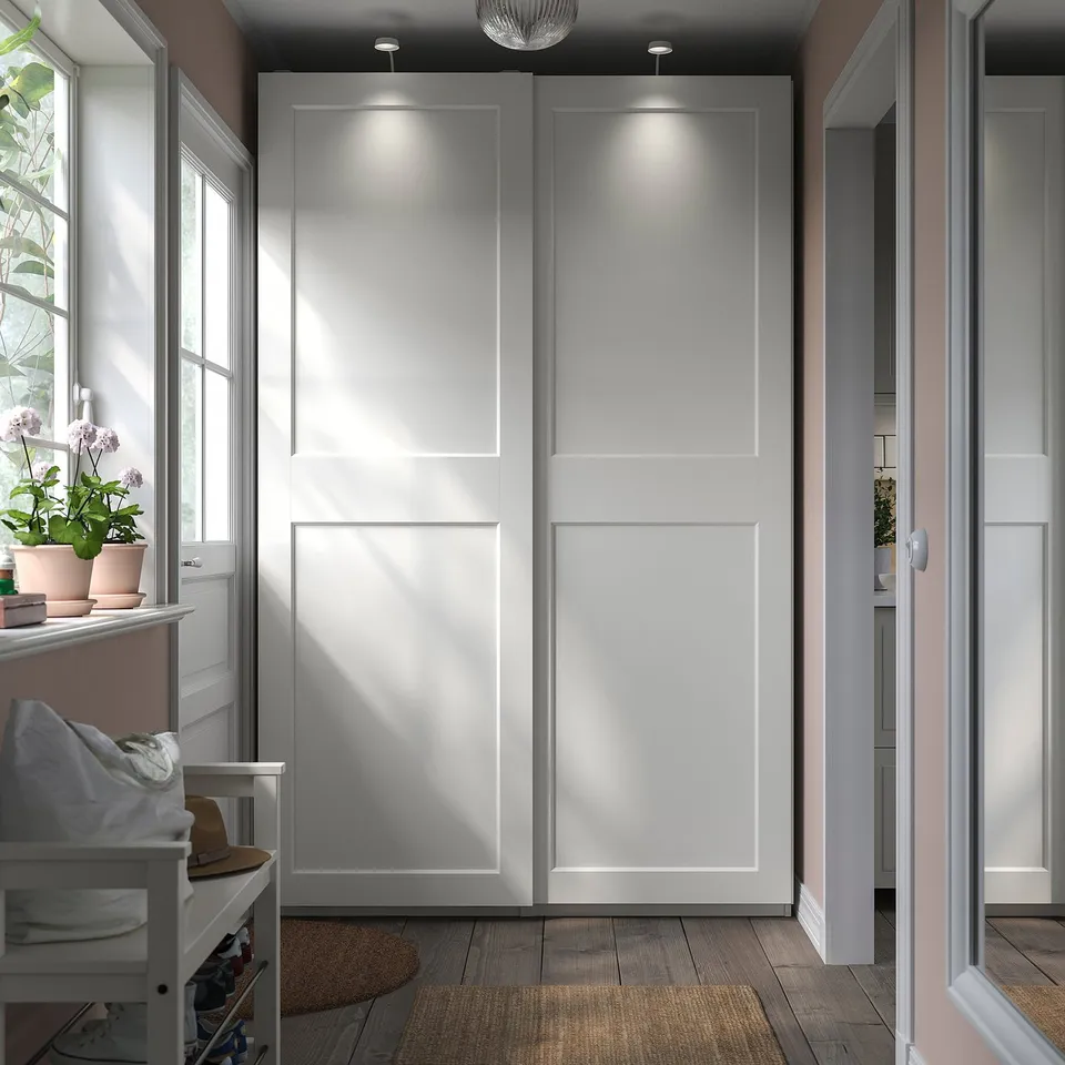 Minimalist hallway with white PAX wardrobe, subtle lighting, wooden floors, and potted plants near windows.