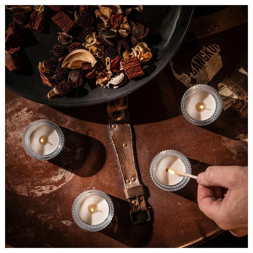 Four lit candles in silver holders on a wooden table, surrounded by dried plant material in a black bowl; person holding a matchstick.