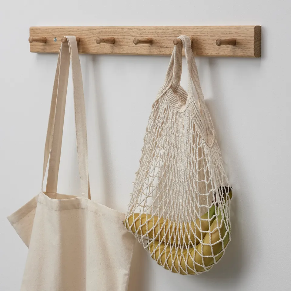 A wooden rack with three knobs holding a canvas bag and a net bag filled with fruits.
