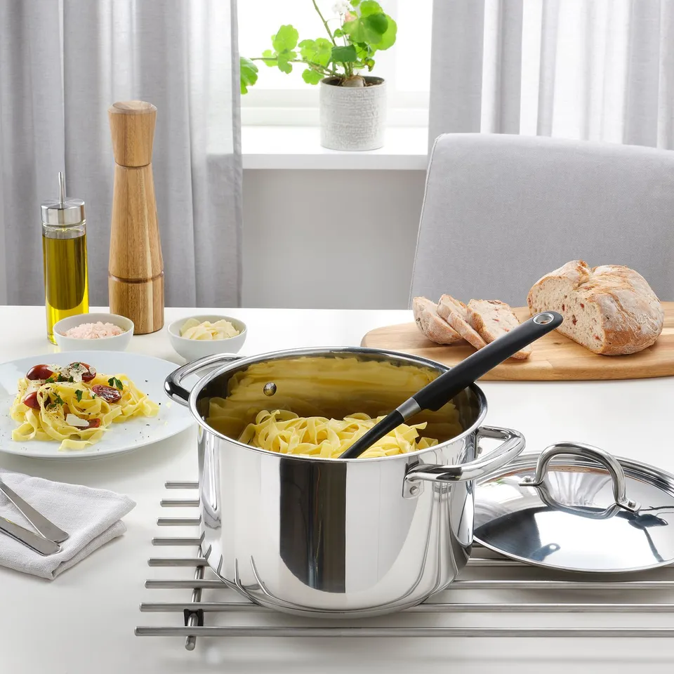 Silver saucepan on stove with pasta, wooden cutting board, bread, olive oil, salt, pepper, and dish with food.