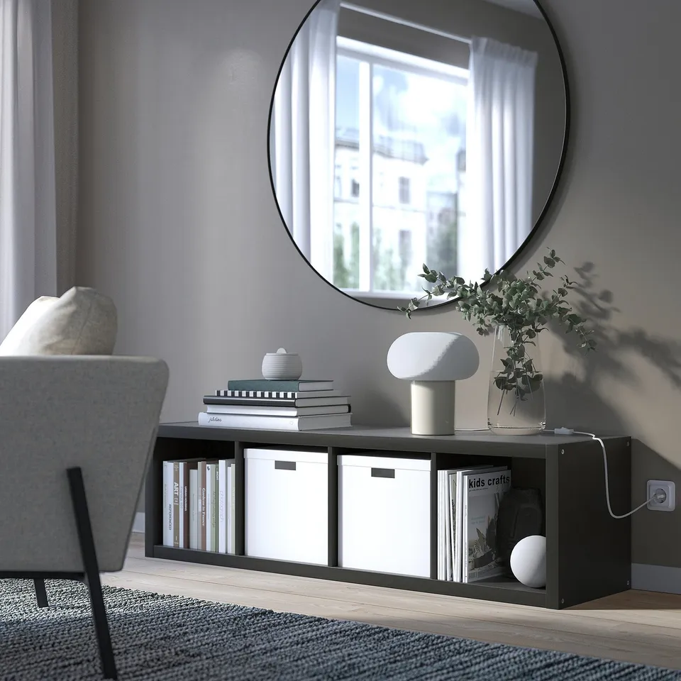 Modern living room with KALLAX black shelf, grey armchair, white lamp, greenery, books, magazines, and round wall mirror reflecting window.