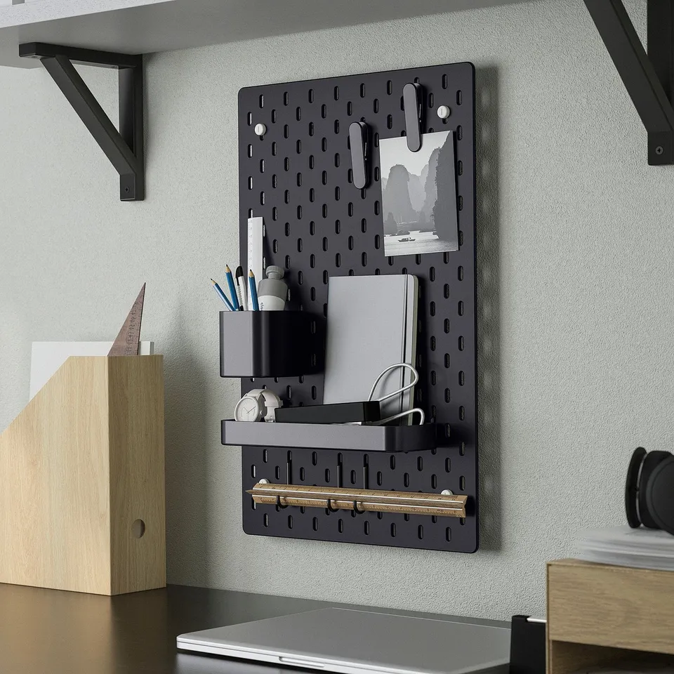 Black SKÅDIS pegboard organiser holds office supplies, notes, and accessories. Mounted on wall with shelf and hooks for storage.
