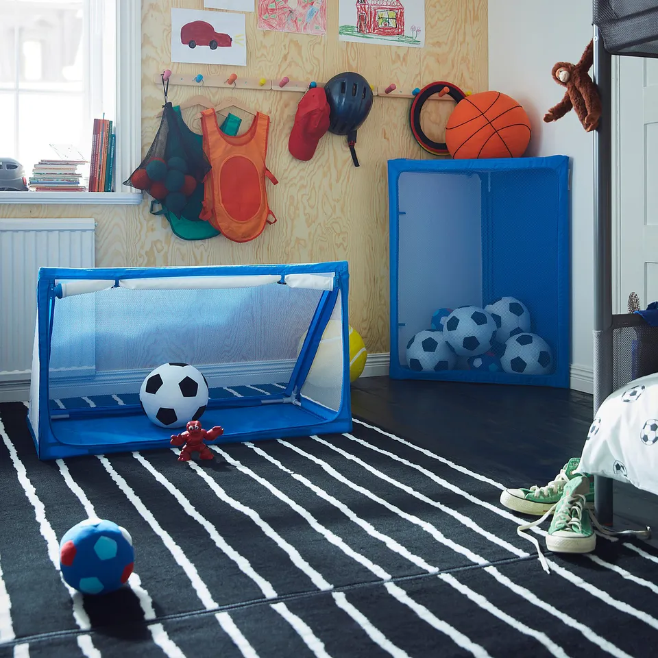 Childrens play area with blue SPORTSLIG ball storage, football net, and various toys. Wood-paneled wall with drawings and sports equipment.