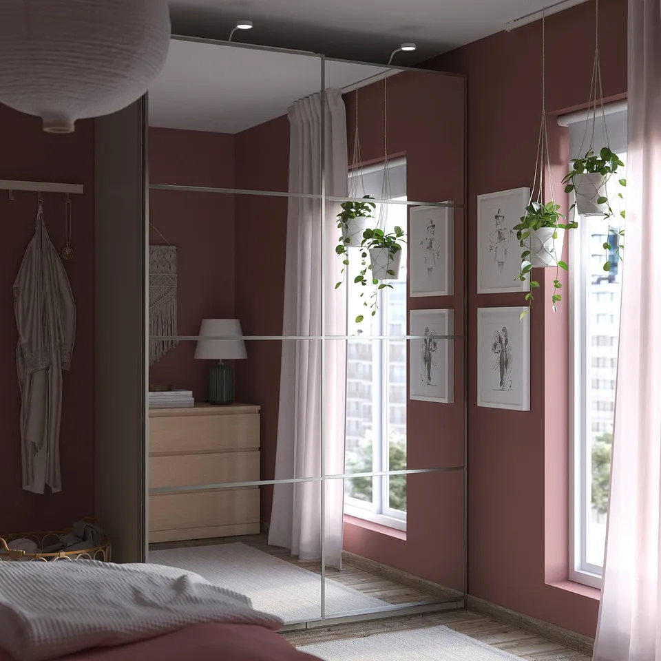 Bright bedroom with brown walls, white bedding, pink curtains, hanging plants, large mirror, and wooden dresser.