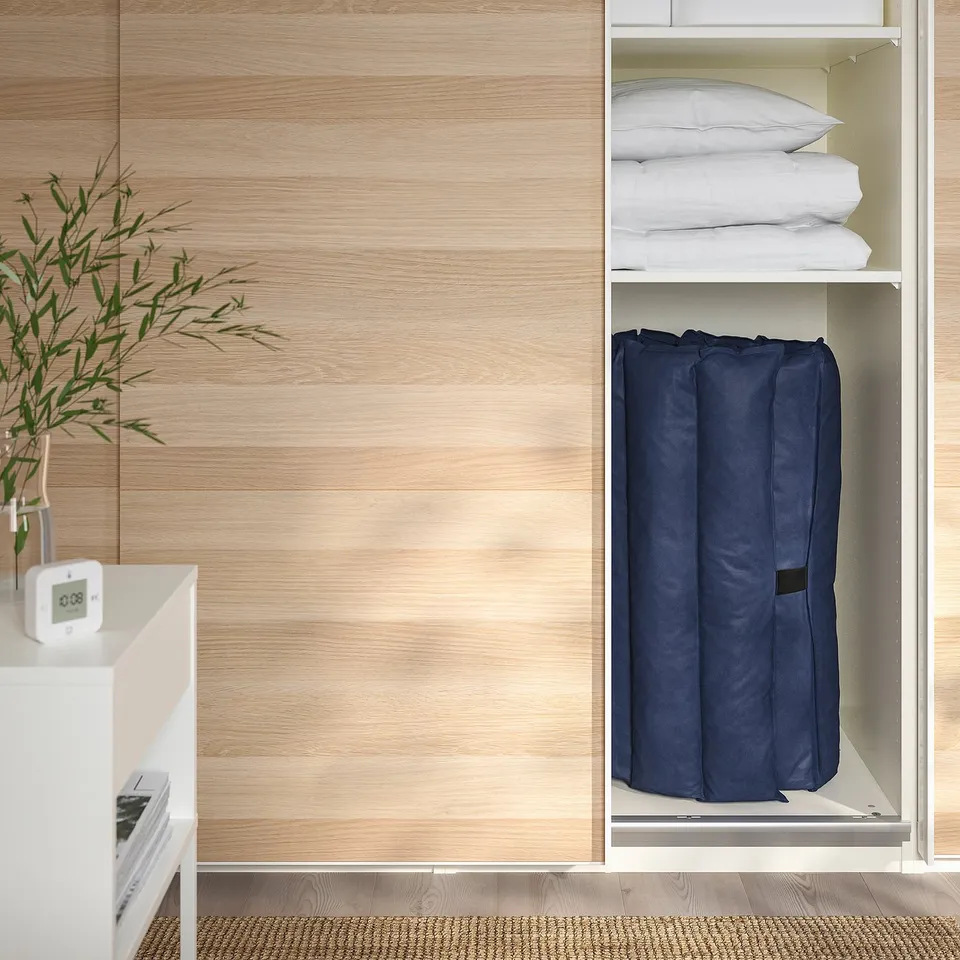 Closet with folded futon mattress and pillows, bedside table with alarm clock and plant.