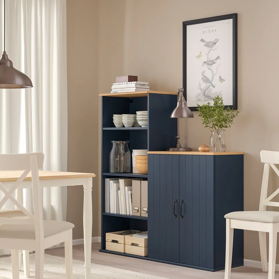 Navy SKRUVBY cabinet/bookcase in dining area with doors, wooden top & decorative items. Adjacent white dining table with four chairs.