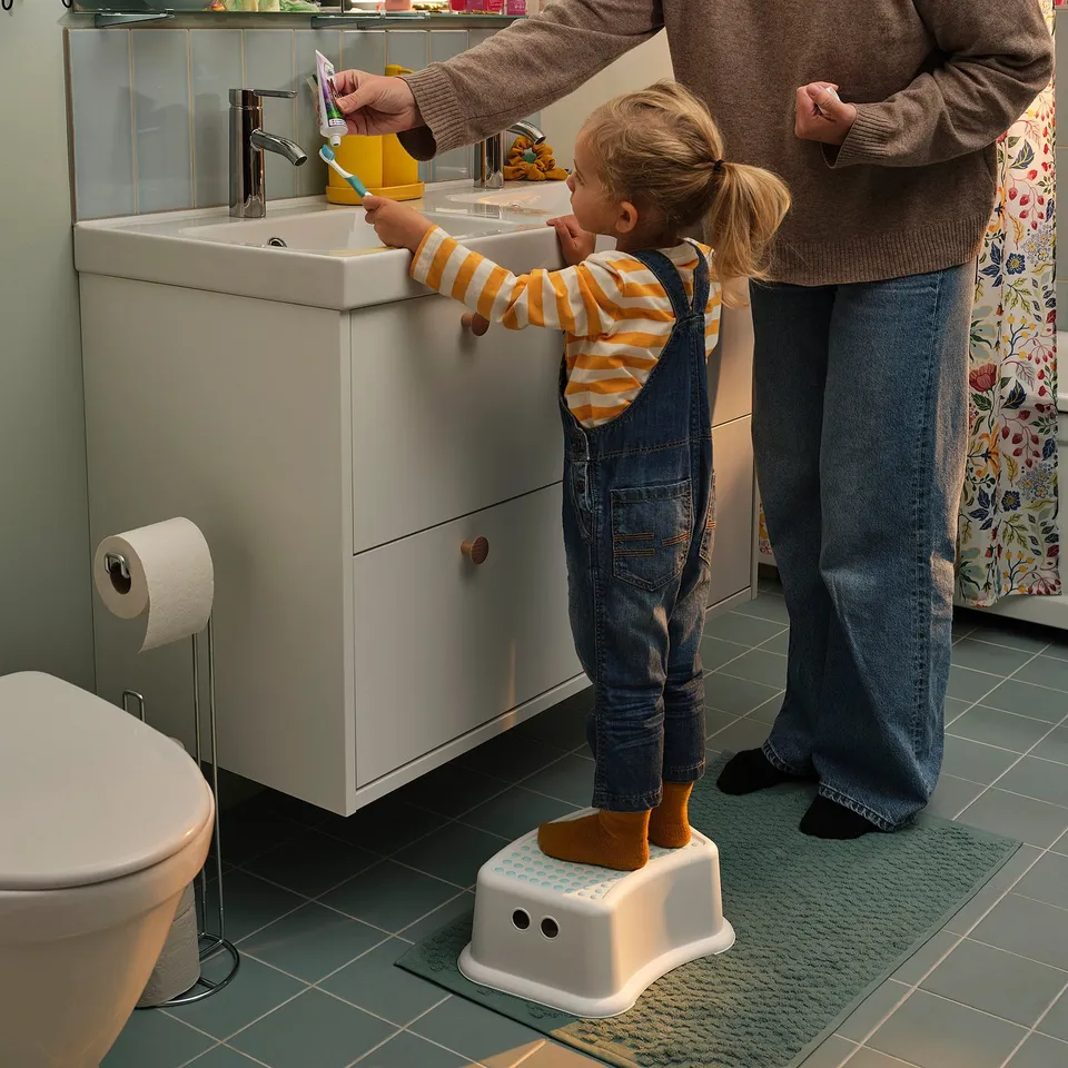 Child stands on white FÖRSIKTIG stool brushing teeth beside adult in bathroom.