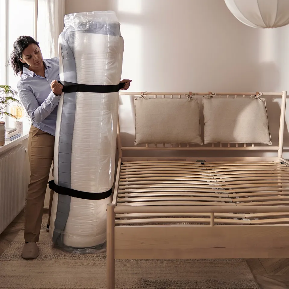 A person unboxes a new rolled mattress next to a wooden bed frame.