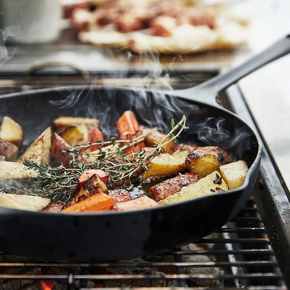 Black skillet with roasted vegetables, grilling. Smoke rises. Background, dish with sauce.
