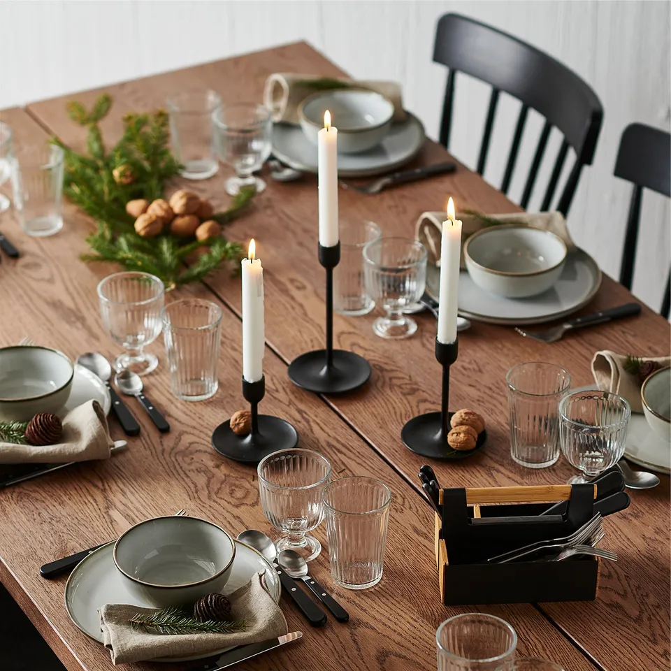 Formal dining table set with grey plates, black cutlery, white candles, clear glasses, green napkins, and pine cones.