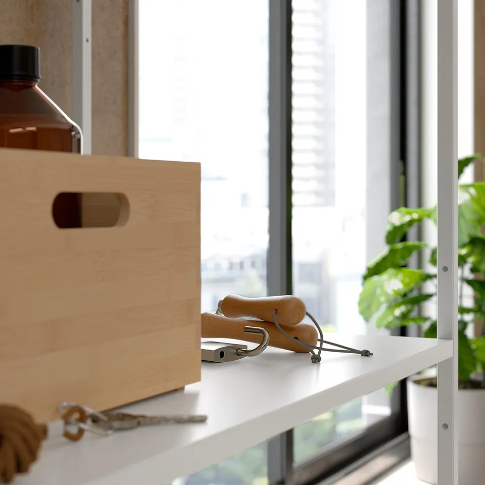 A white metal shelf holding wooden box, exercise equipment, and keys, near plants by a window.