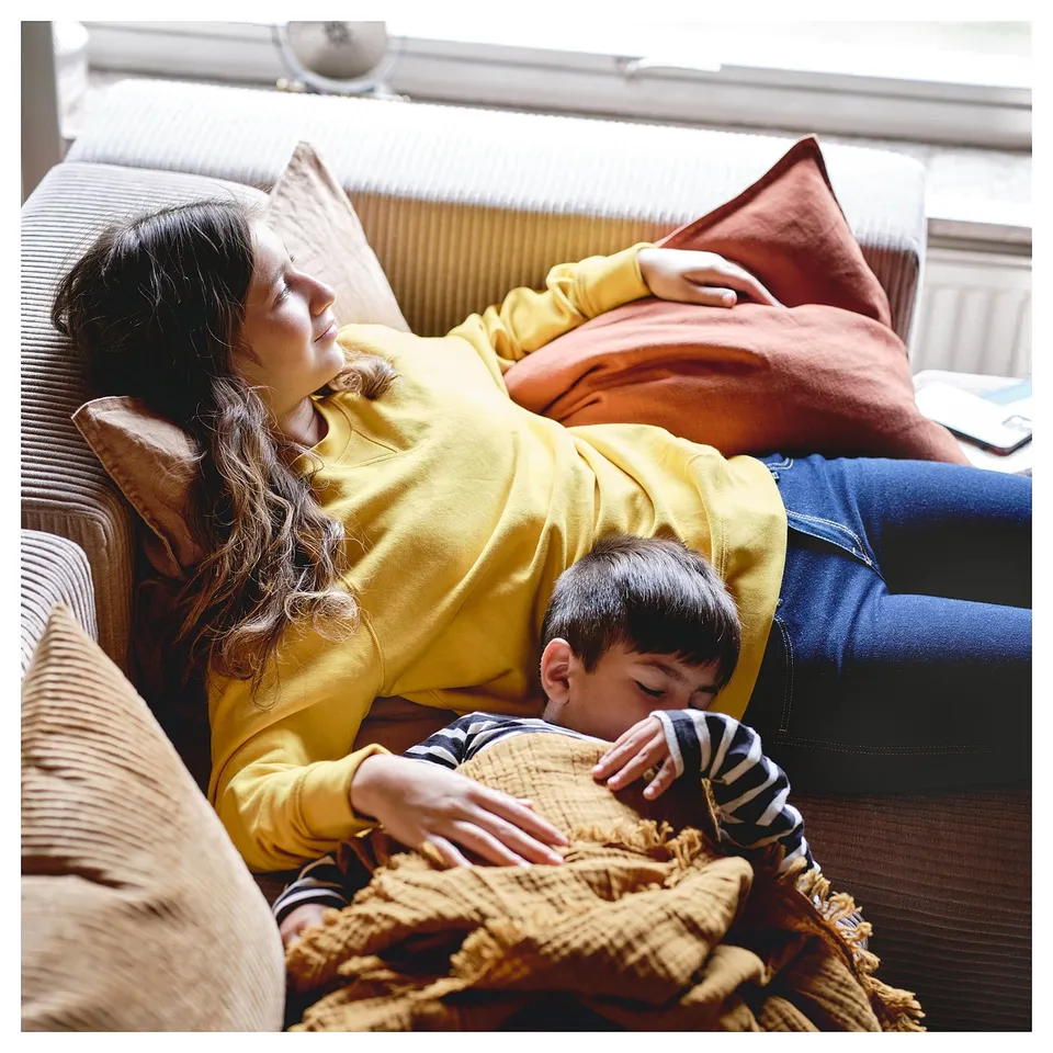 A person and a child relax on a yellow JÄTTEBO modular sofa, snuggling under a blanket.