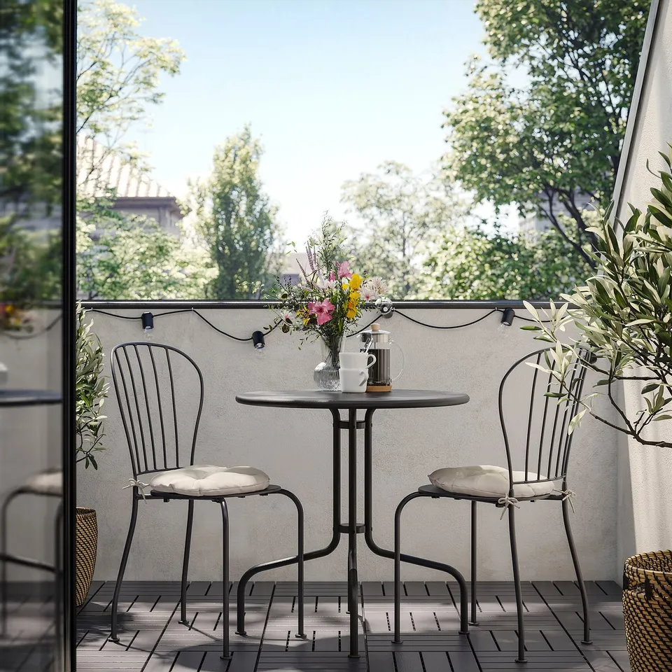 Small black outdoor table setup with two chairs, cushioned seats, a vase of flowers, and string lights on a balcony.