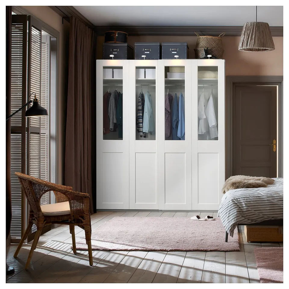 Modern bedroom with glass-front PAX wardrobe, chair, rug, and striped bed. Wood floor, beige walls, large window.