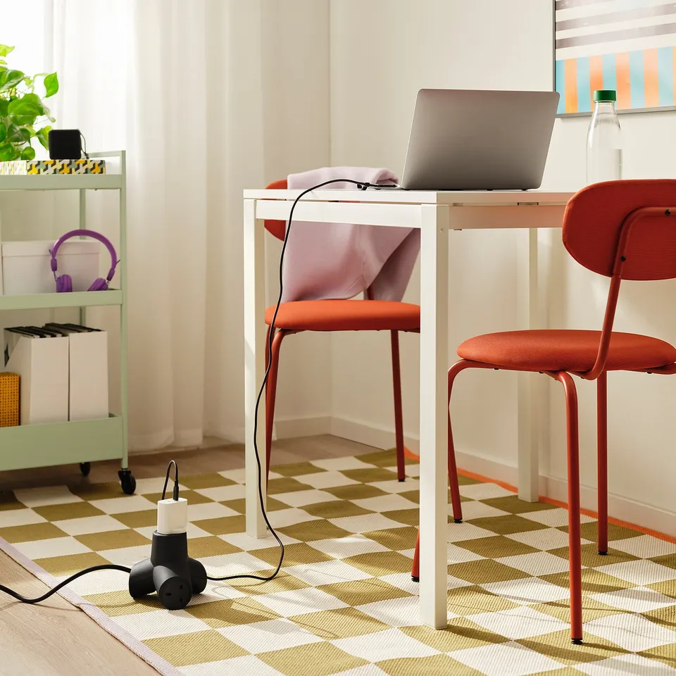 White desk with laptop, chair, purple headphones, green shelving, and a black, 4-way extension cord on the floor. Chequered rug.