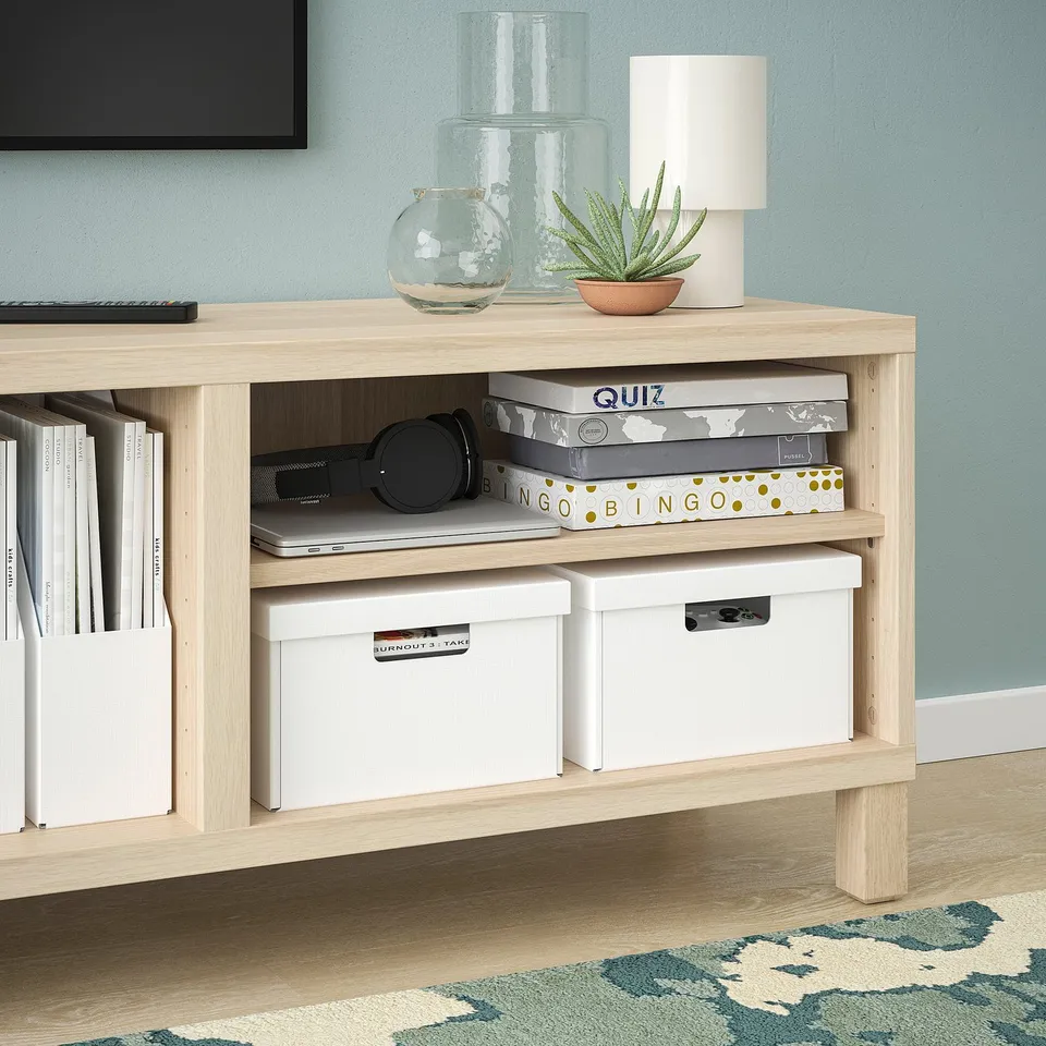 Wooden sideboard with shelves holding books, game boxes, and storage bins. A lamp, plants, and decorative vases on top.