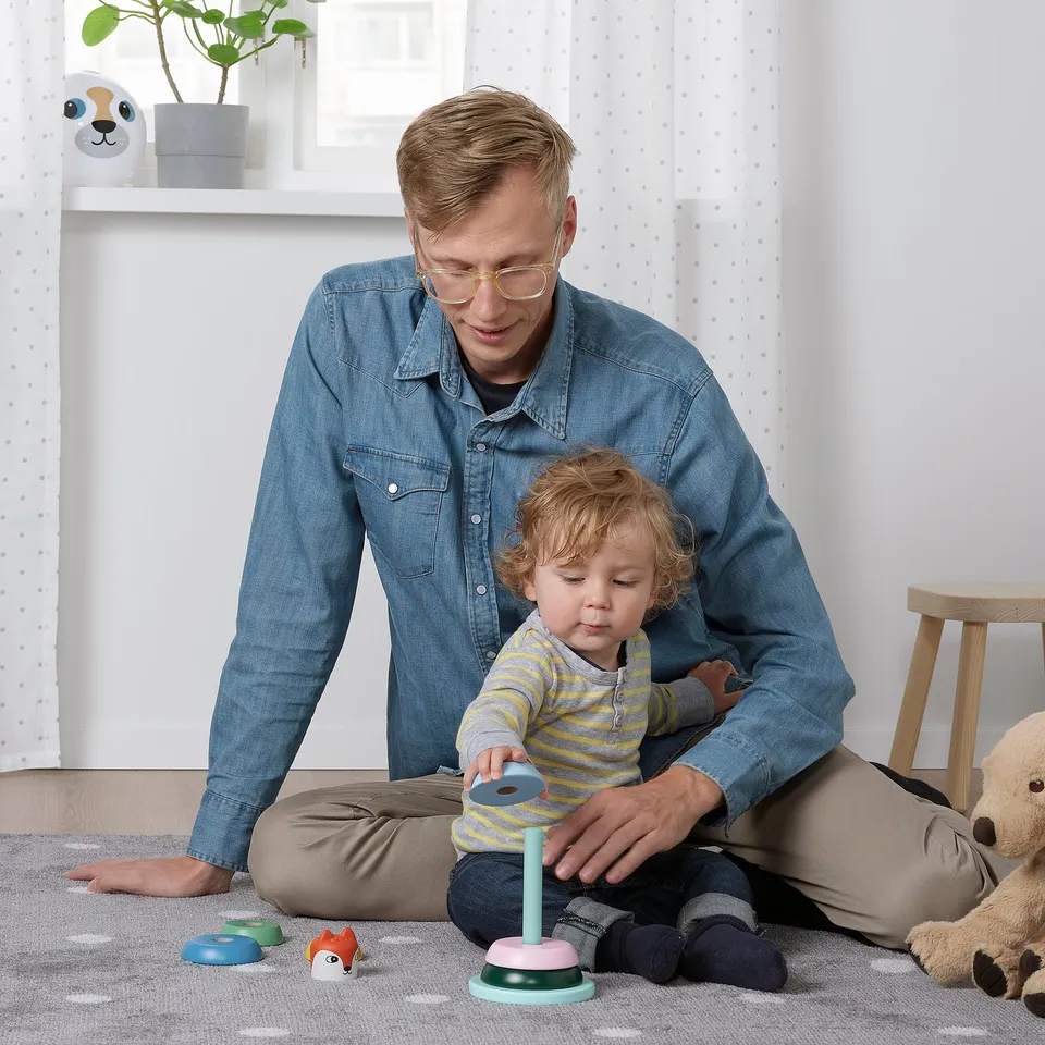 A person and toddler play with UPPSTÅ stacking toy, stacking coloured rings on a soft stick base.