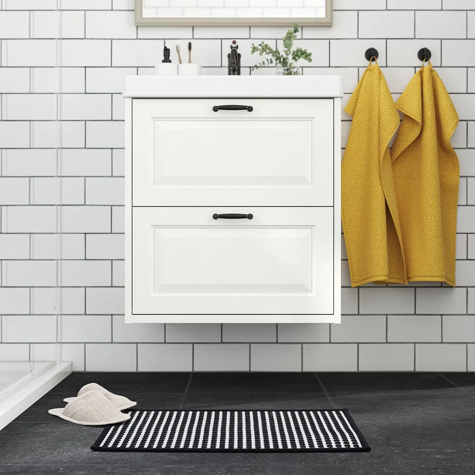 Modern bathroom with white cabinet and tiles, mustard towels on black hooks, black & white bath mat.