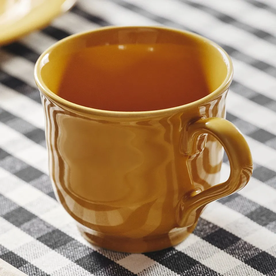Close-up of a glossy, mustard-yellow mug on a checked tablecloth. Mug has a smooth handle and reflects light, showcasing its high-quality texture.