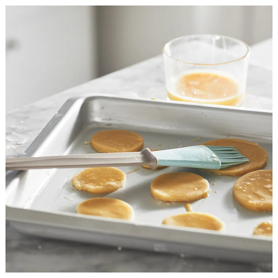 Silver baking tray with several round cookies being brushed with a light blue silicone brush dipped in a yellow liquid in a clear glass nearby.