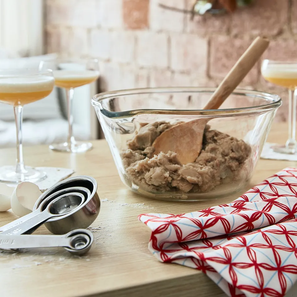 Wooden spoon in glass bowl of dough on table with measuring cups, wine glasses, cloth napkin, brick wall background.