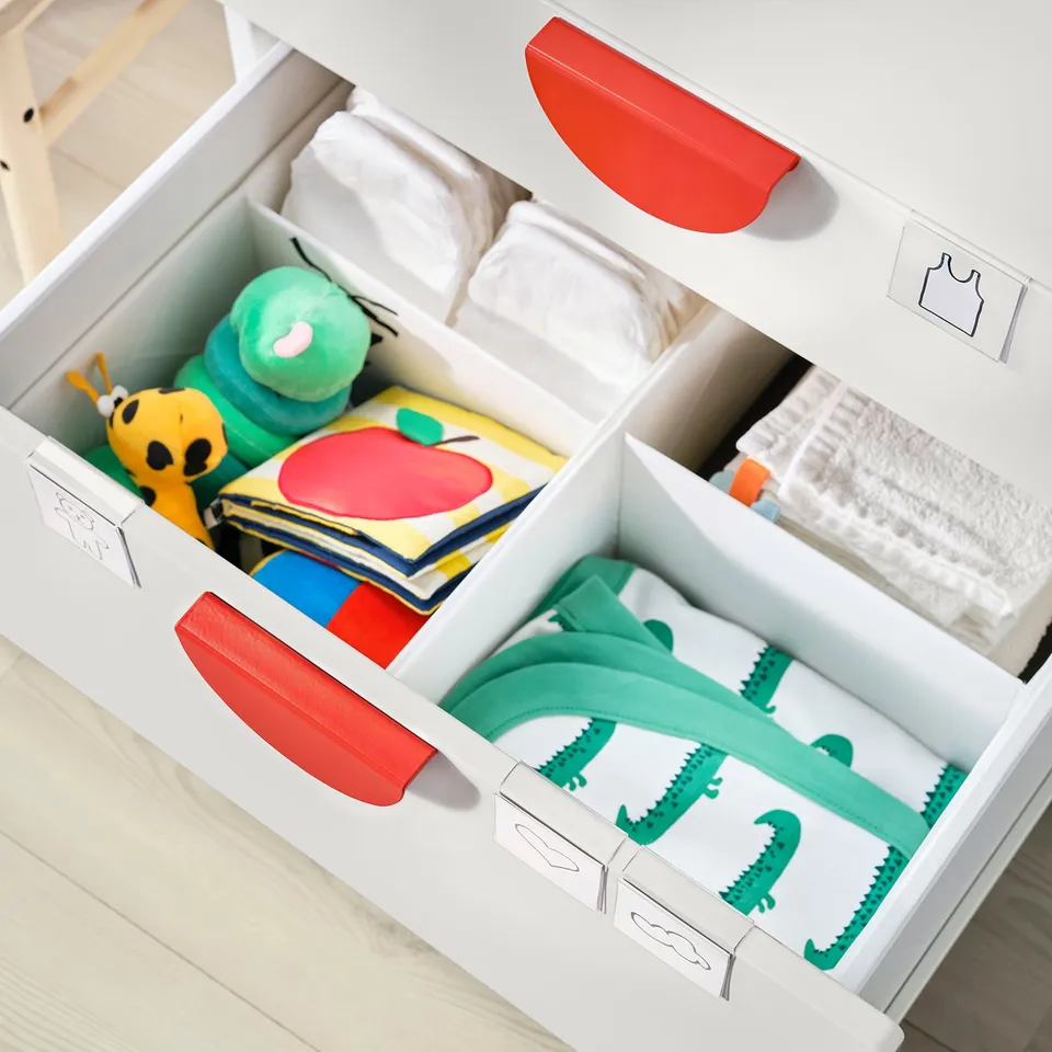 White nursery organiser with red handles, storing baby clothes and toys, including green dinosaur pyjamas.