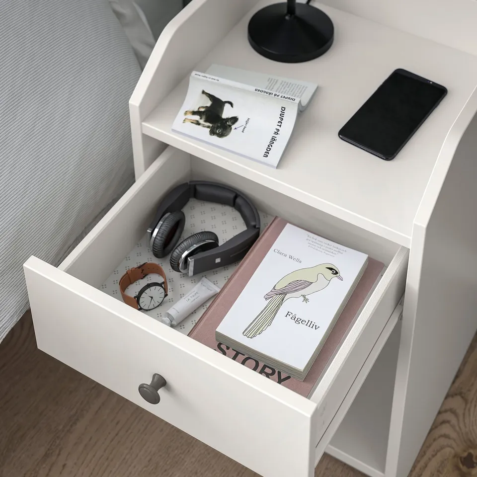 White bedside table with open shelf, drawer, and storage area. Contains books, phone, and headphones.