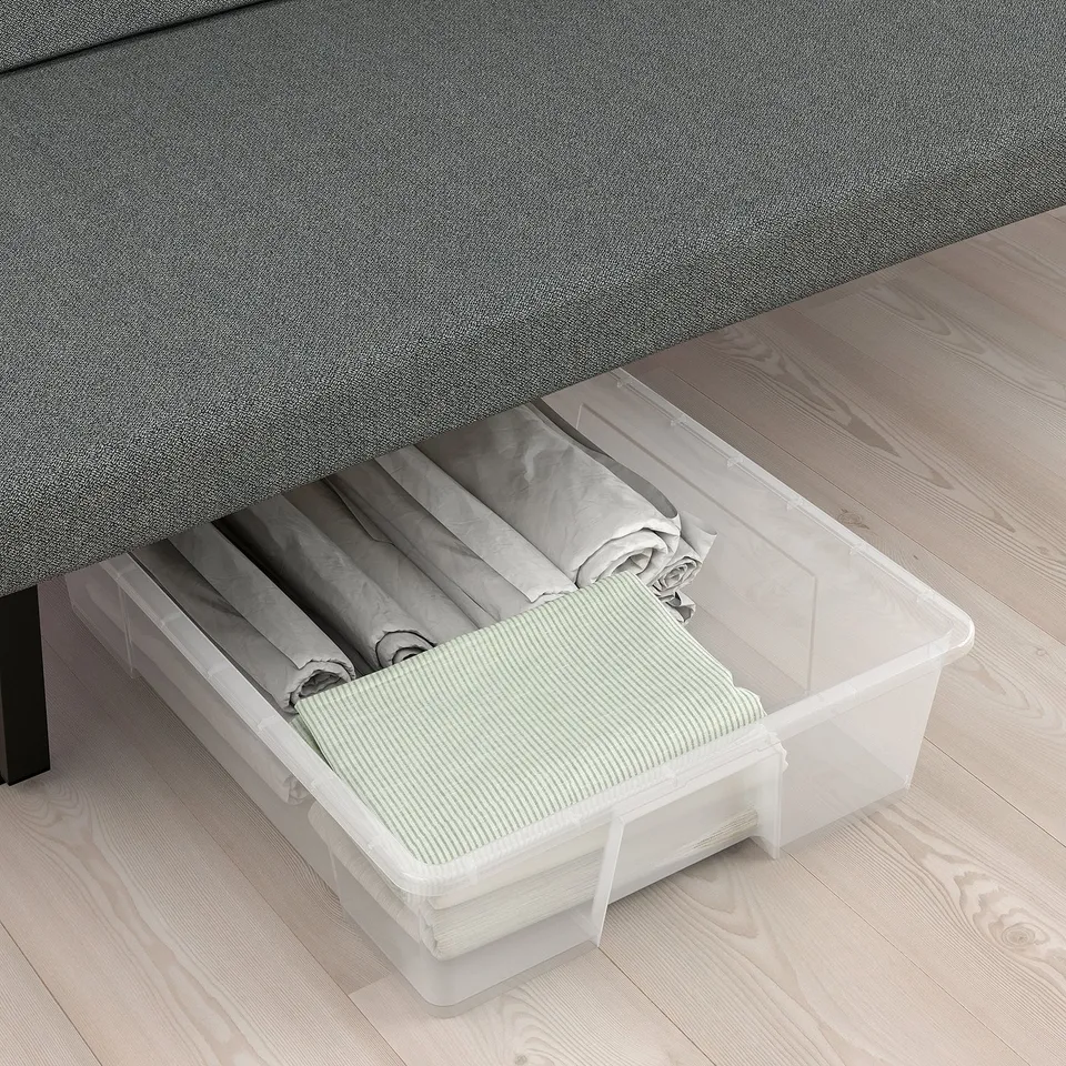 Clear plastic storage box under grey sofa, holding folded blankets and striped pillowcases on wooden floor.