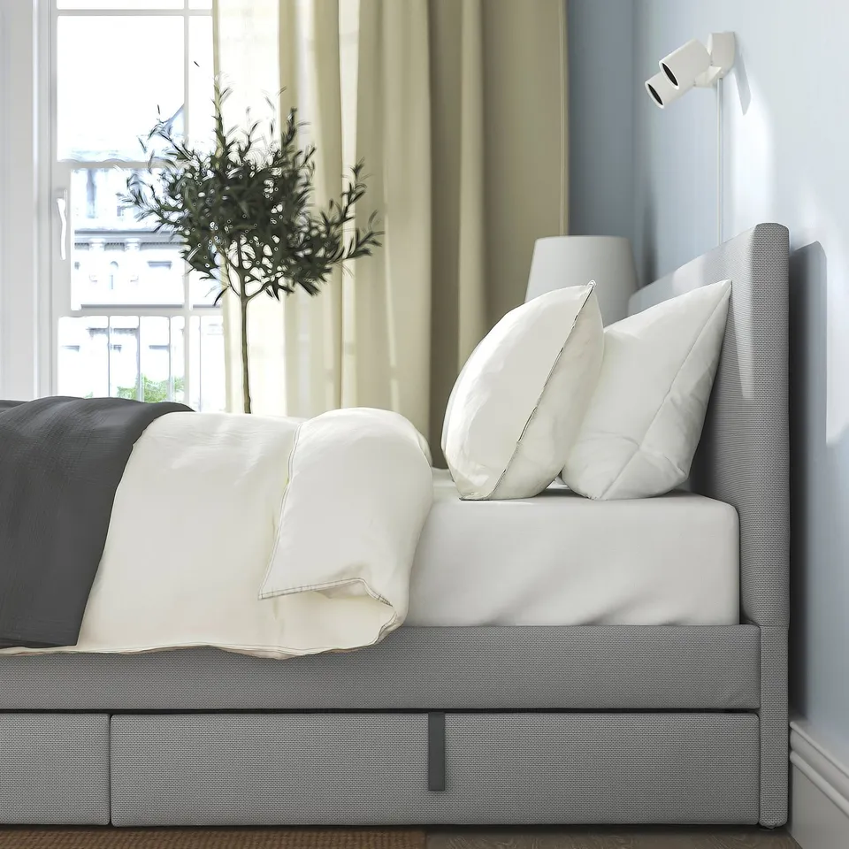 Modern bed with grey upholstery, white pillows, and a padded headboard. A small tree in a vase is placed beside it.