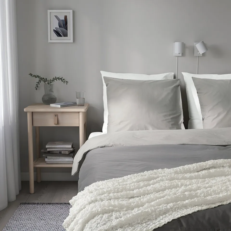 Modern bedroom with white bedding, fluffy blanket, wooden bedside table, books, lamp, and plant.