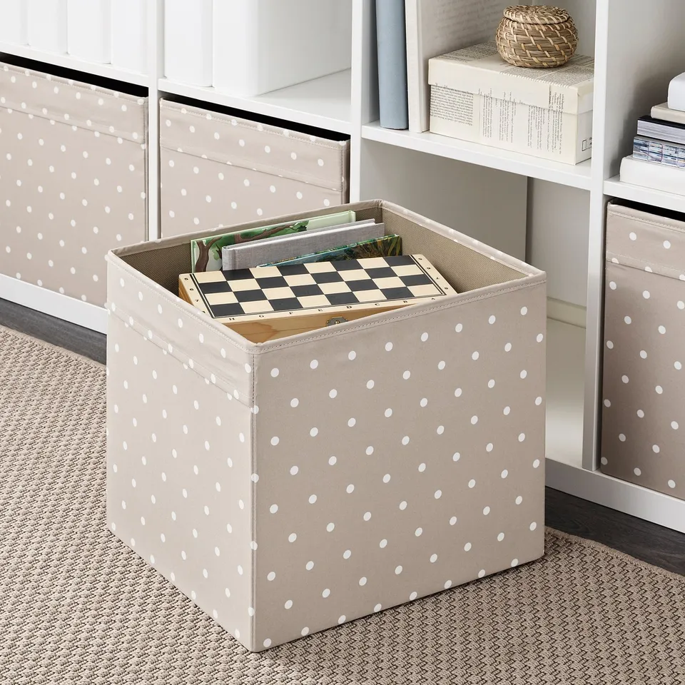 Fabric storage box with white polka dots sits on a shelf, holding game boards and papers. Designed for kallax shelving, it organises items neatly.