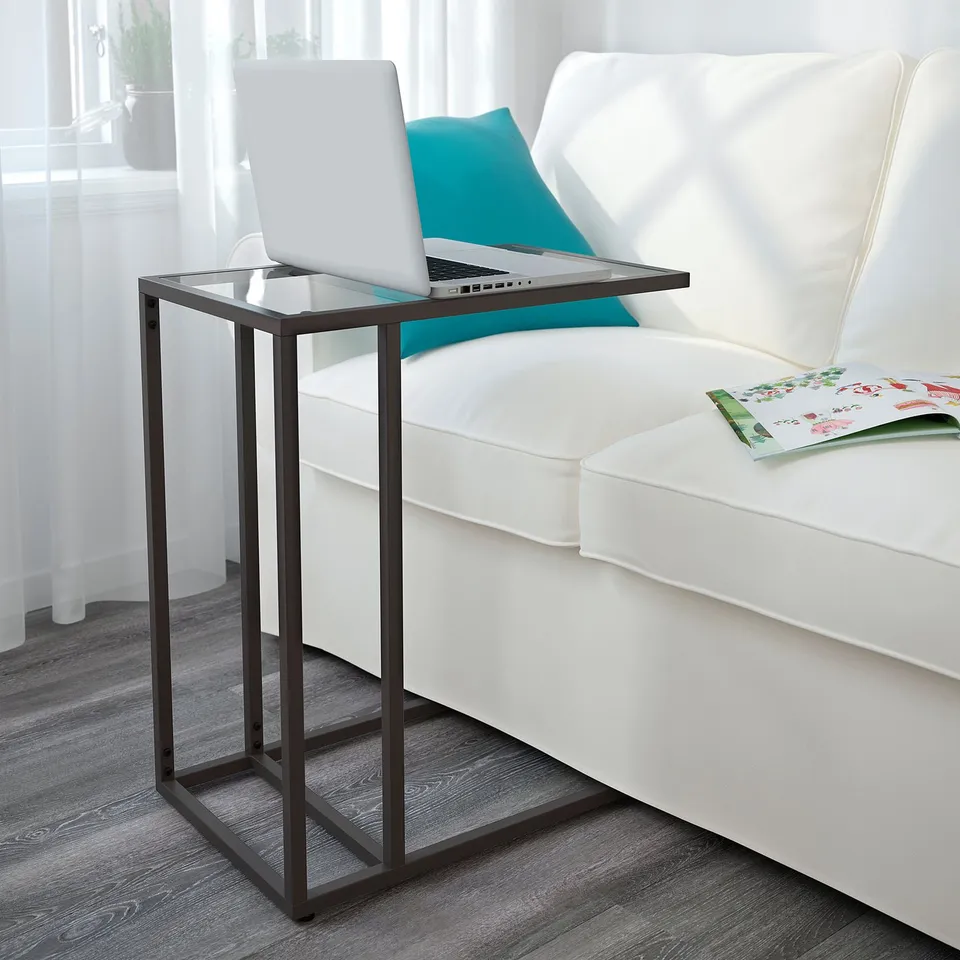 Small glass-top side table next to white couch, holding a laptop and a book.