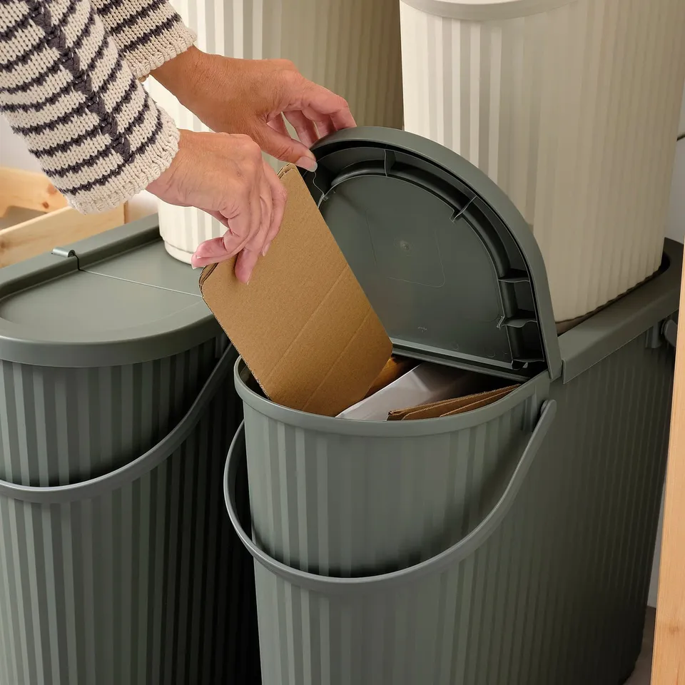 Person placing corrugated cardboard into a grey recycling bin with a lid. The bin has a handle for easy movement.
