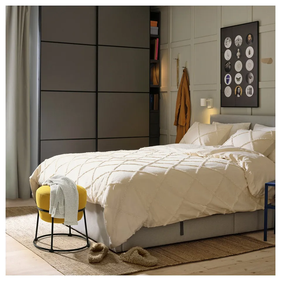 Bedroom with unmade bed, white bedding, yellow stool, furry slippers, wall art, coat hooks, and wooden floor.