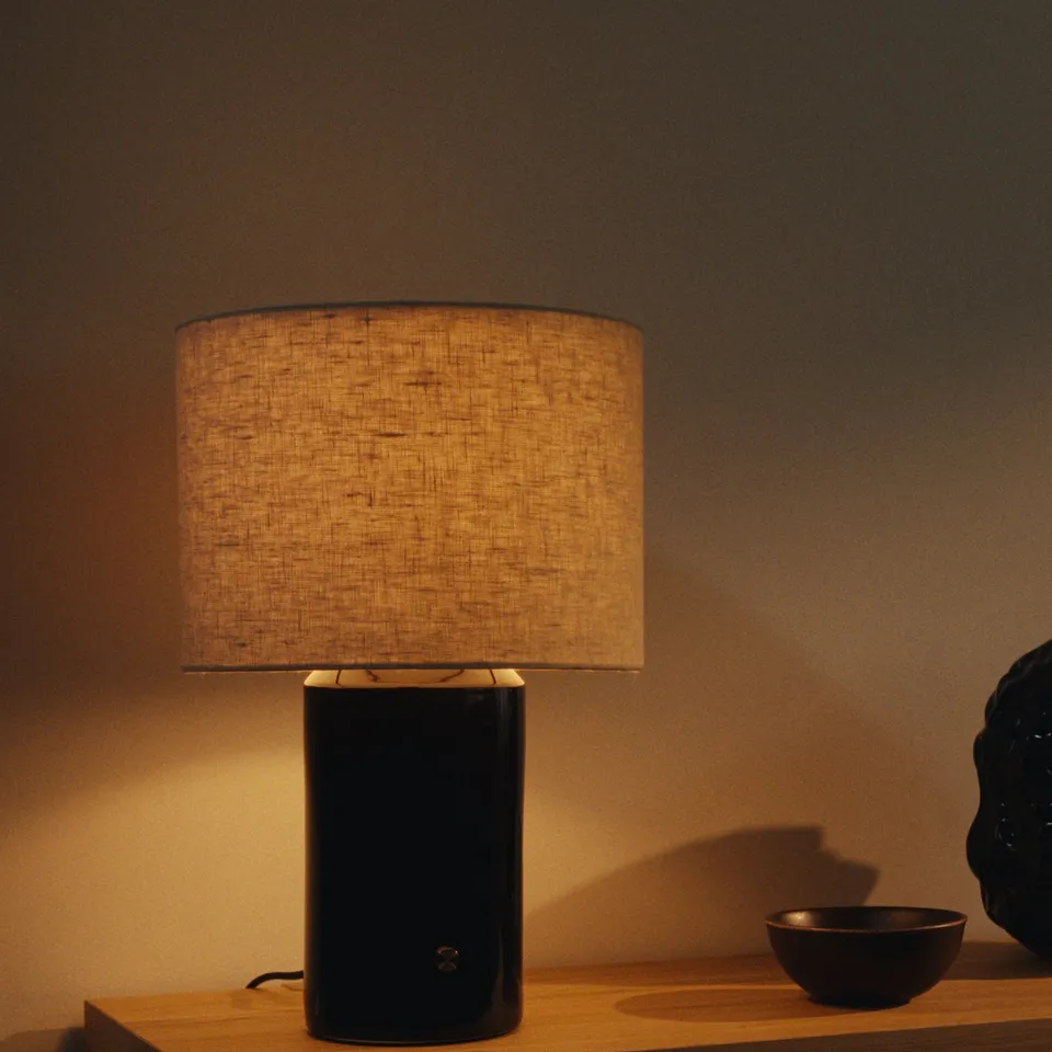A HALGATT lamp with a beige linen shade sits on a wooden surface, casting a warm glow. The base is dark, ceramic with a brass dimmer knob.