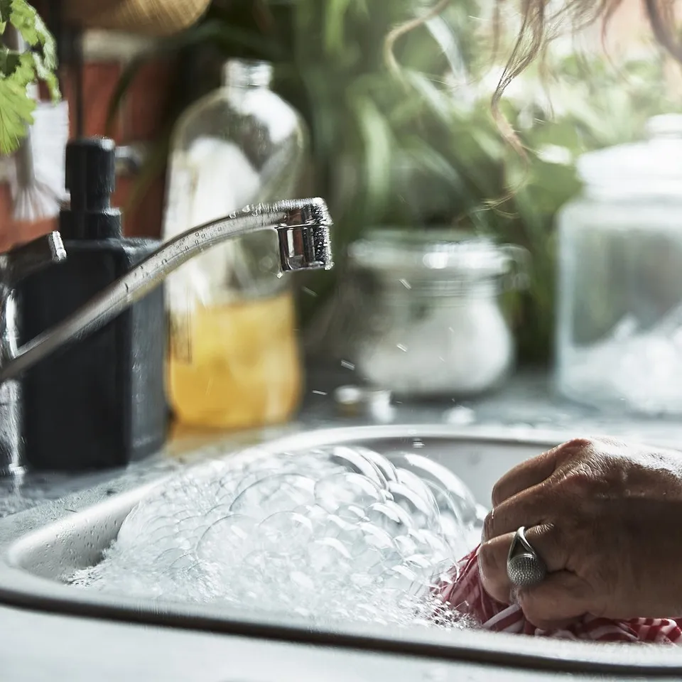 Person washing produce in outdoor sink. Stainless steel tap, soapy water, green plants in background.