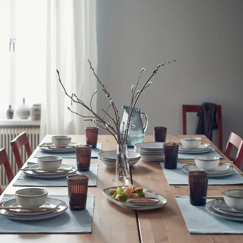 Dining table with GLADELIG bowls, glasses, blue placemats, and budding branch centrepiece. Sandy-glazed bowls, wooden chairs, curtains.