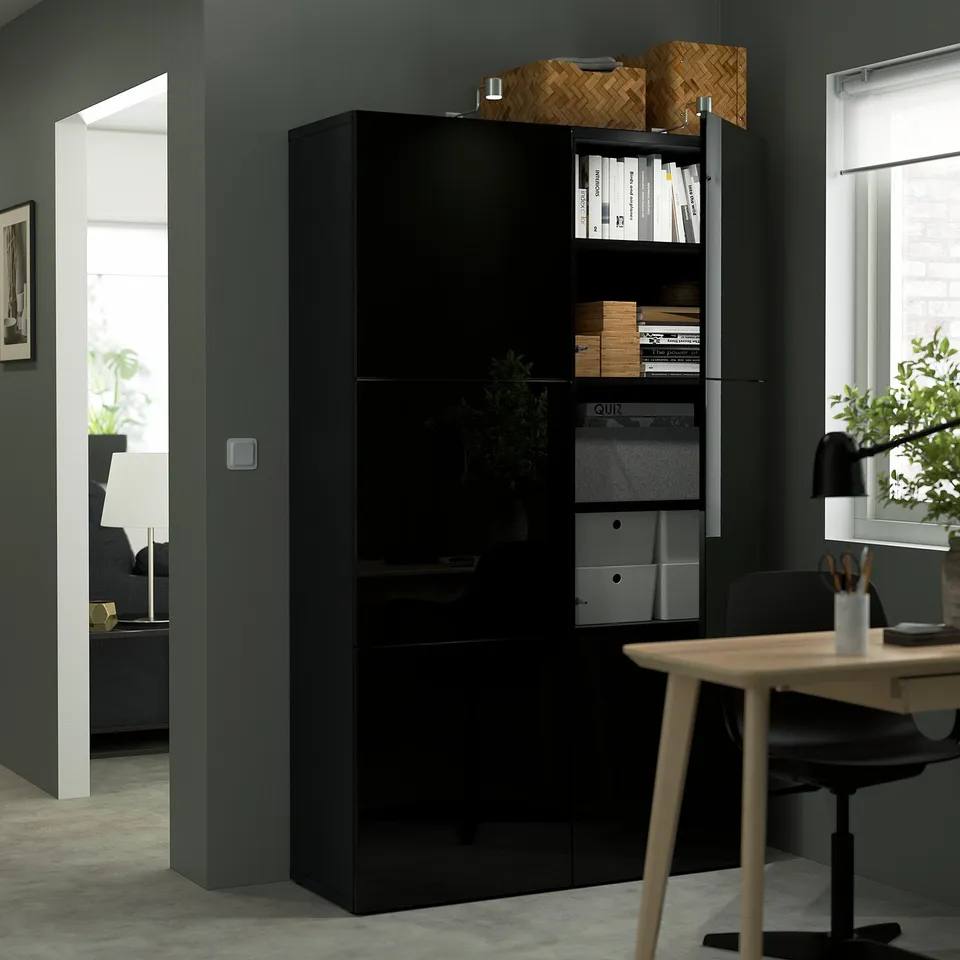 Black BESTÅ storage unit with open shelves holding files and boxes, against dark green wall. Next to wooden table with chair.