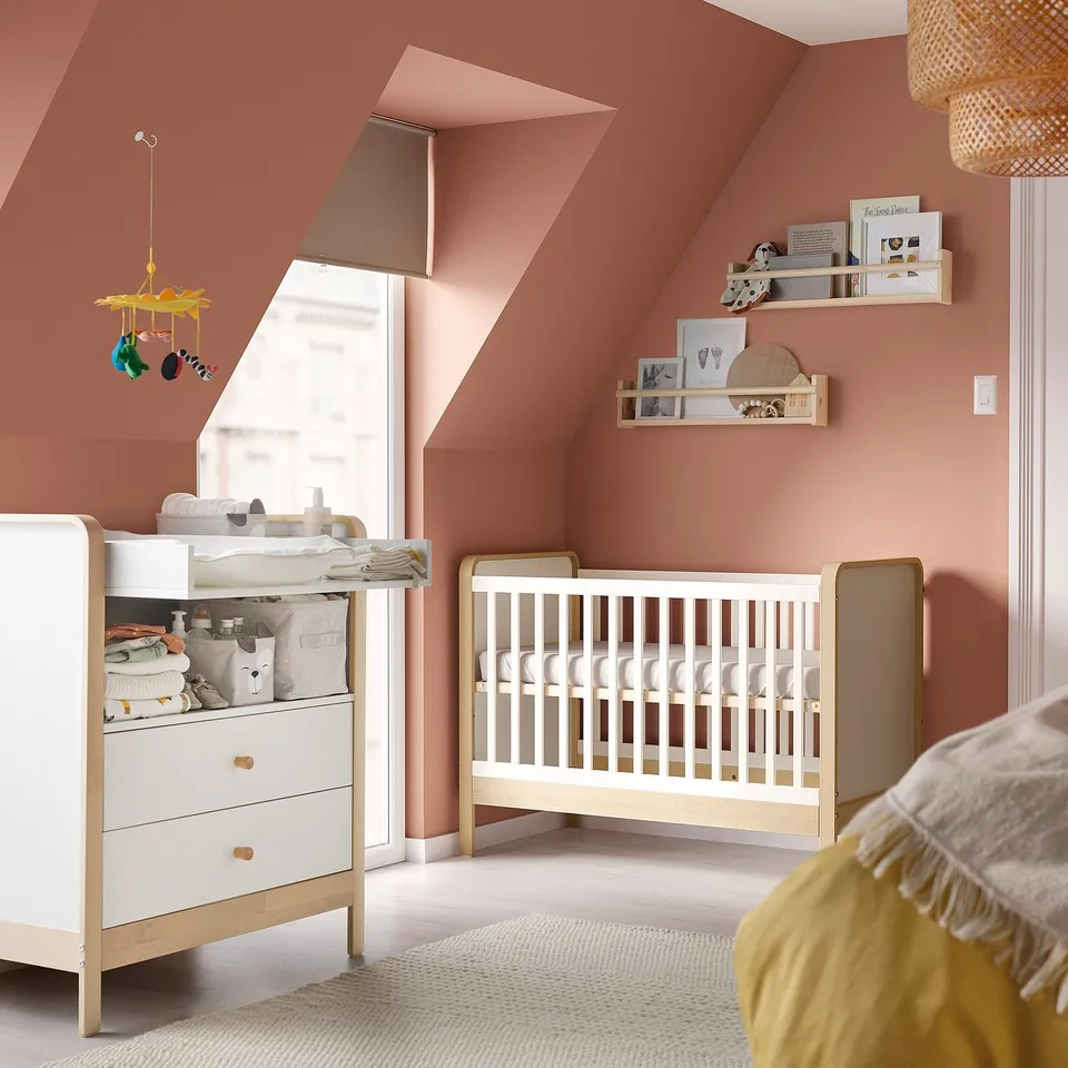 Modern nursery with pink walls, white cot, changing table, and hanging mobile.