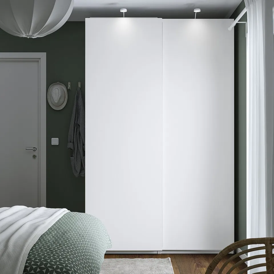 Minimalist white wardrobe in green bedroom. Doors closed, hat & towel on hooks, bed with blanket, chair partly visible.